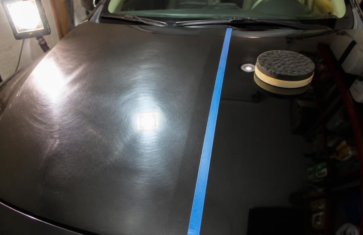 Test spot on a dark car hood divided by masking tape, showing hazy uncorrected paint next to a polished section with sharper reflections